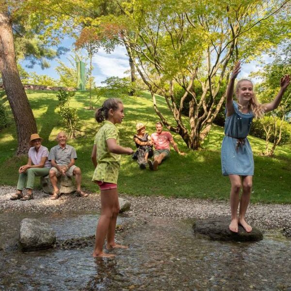 Zwei Mädchen spielen auf Steinen in einem seichten Bach, während vier Erwachsene an einem sonnigen Tag auf der Wiese in der Nähe sitzen, umgeben von Bäumen und Grünpflanzen.