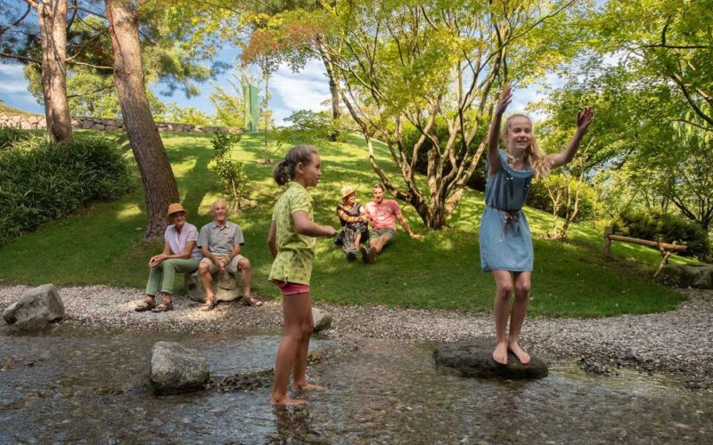 Zwei Mädchen spielen auf Steinen in einem seichten Bach, während vier Erwachsene an einem sonnigen Tag auf der Wiese in der Nähe sitzen, umgeben von Bäumen und Grünpflanzen.