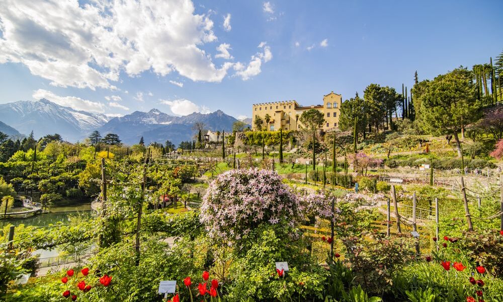 Ein großer formaler Garten mit blühenden Blumen und Pflanzen, Bäumen und einer gelben Villa im Hintergrund, umgeben von Bergen unter einem teilweise bewölkten Himmel.