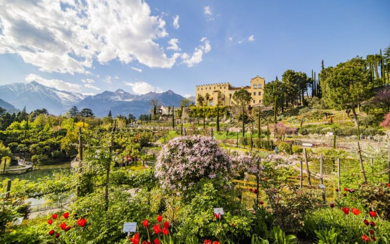 Ein großer formaler Garten mit blühenden Blumen und Pflanzen, Bäumen und einer gelben Villa im Hintergrund, umgeben von Bergen unter einem teilweise bewölkten Himmel.