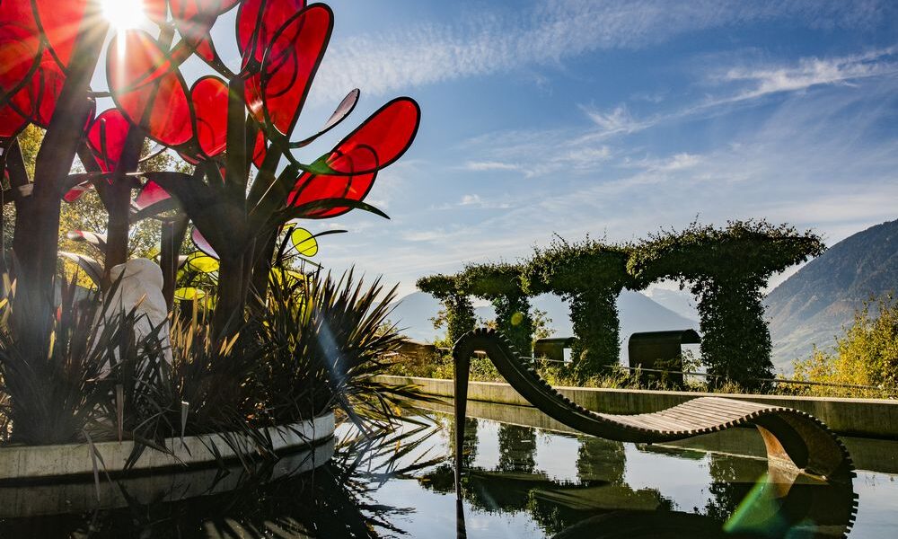 Eine moderne Skulptur mit großen roten Glasblättern steht neben einer geschwungenen Holzbank an einem spiegelnden Teich, mit Bergen und gestutzten Hecken im Hintergrund unter einem strahlenden Himmel.