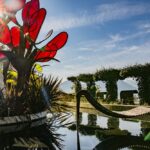Eine moderne Skulptur mit großen roten Glasblättern steht neben einer geschwungenen Holzbank an einem spiegelnden Teich, mit Bergen und gestutzten Hecken im Hintergrund unter einem strahlenden Himmel.