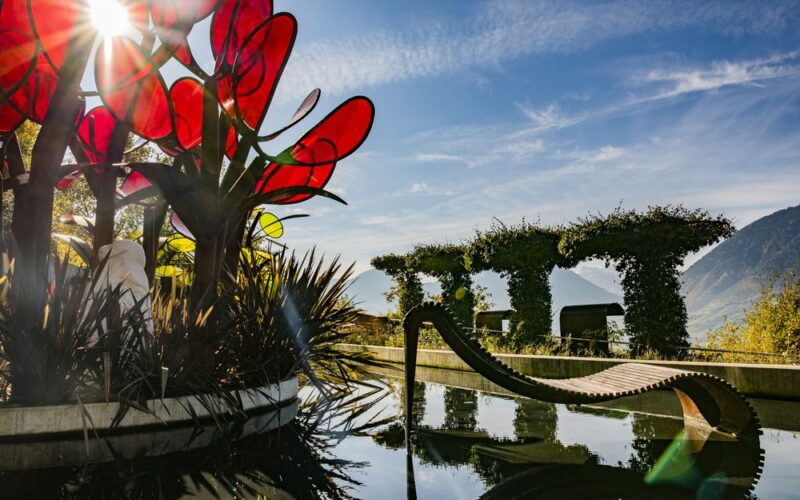 Eine moderne Skulptur mit großen roten Glasblättern steht neben einer geschwungenen Holzbank an einem spiegelnden Teich, mit Bergen und gestutzten Hecken im Hintergrund unter einem strahlenden Himmel.