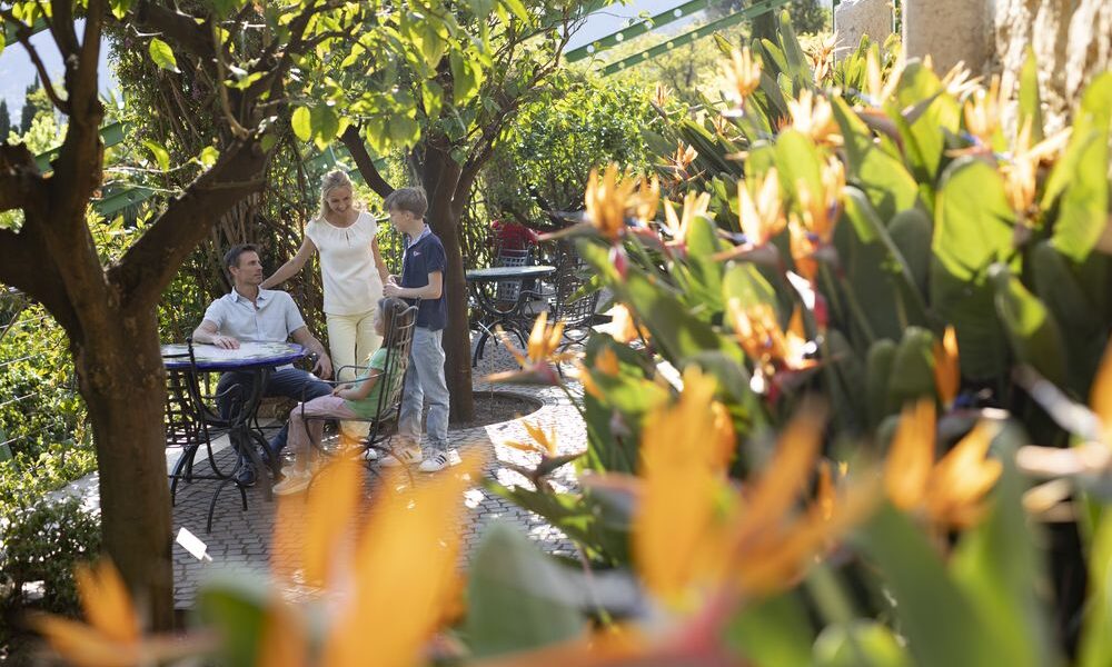 Eine vierköpfige Familie sitzt und steht um einen Tisch in einem Gartencafé, umgeben von üppigem Grün und Paradiesvogelblumen.