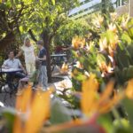 Eine vierköpfige Familie sitzt und steht um einen Tisch in einem Gartencafé, umgeben von üppigem Grün und Paradiesvogelblumen.