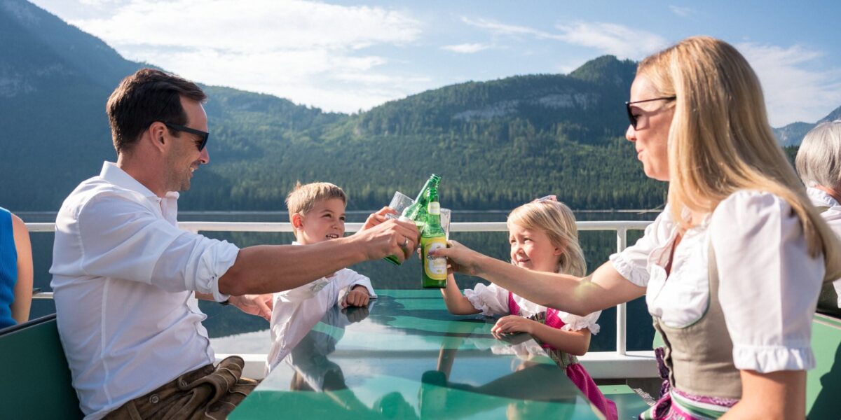 Eine vierköpfige, traditionell gekleidete Familie sitzt an einem Glastisch auf einem Boot und stößt mit Bergen und einem See im Hintergrund an.