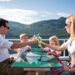 Eine vierköpfige, traditionell gekleidete Familie sitzt an einem Glastisch auf einem Boot und stößt mit Bergen und einem See im Hintergrund an.