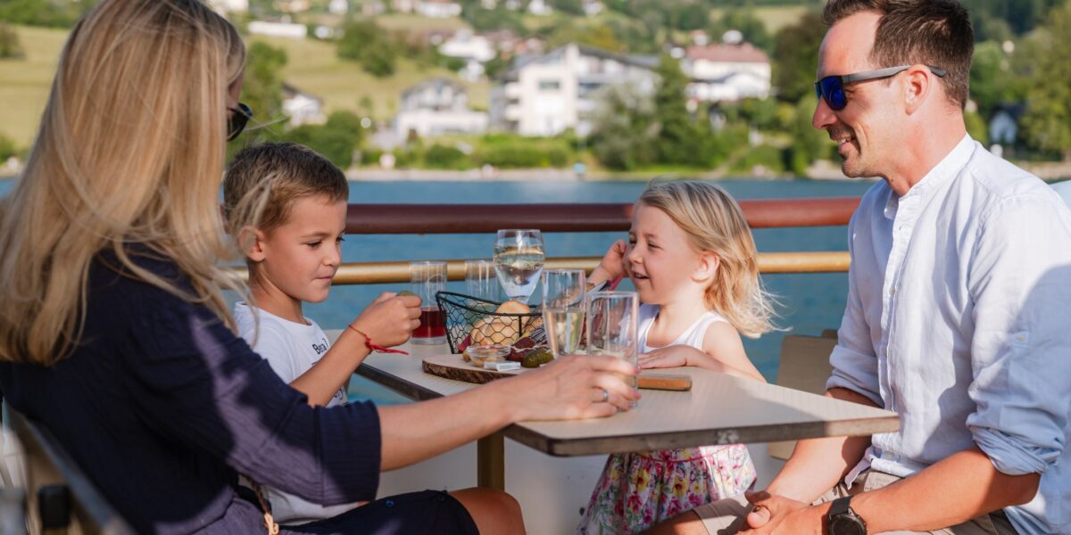 Eine vierköpfige Familie sitzt an einem sonnigen Tag an einem Tisch im Freien und genießt Getränke und Snacks vor einer malerischen Landschaft im Hintergrund.