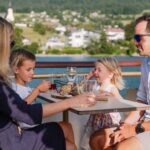 Eine vierköpfige Familie sitzt an einem sonnigen Tag an einem Tisch im Freien und genießt Getränke und Snacks vor einer malerischen Landschaft im Hintergrund.