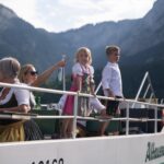 Eine Gruppe von Menschen, darunter auch Kinder in traditioneller Kleidung, fährt auf einem Boot mit Bergen im Hintergrund.