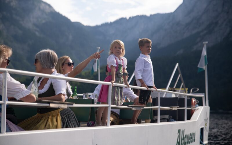 Eine Gruppe von Menschen, darunter auch Kinder in traditioneller Kleidung, fährt auf einem Boot mit Bergen im Hintergrund.