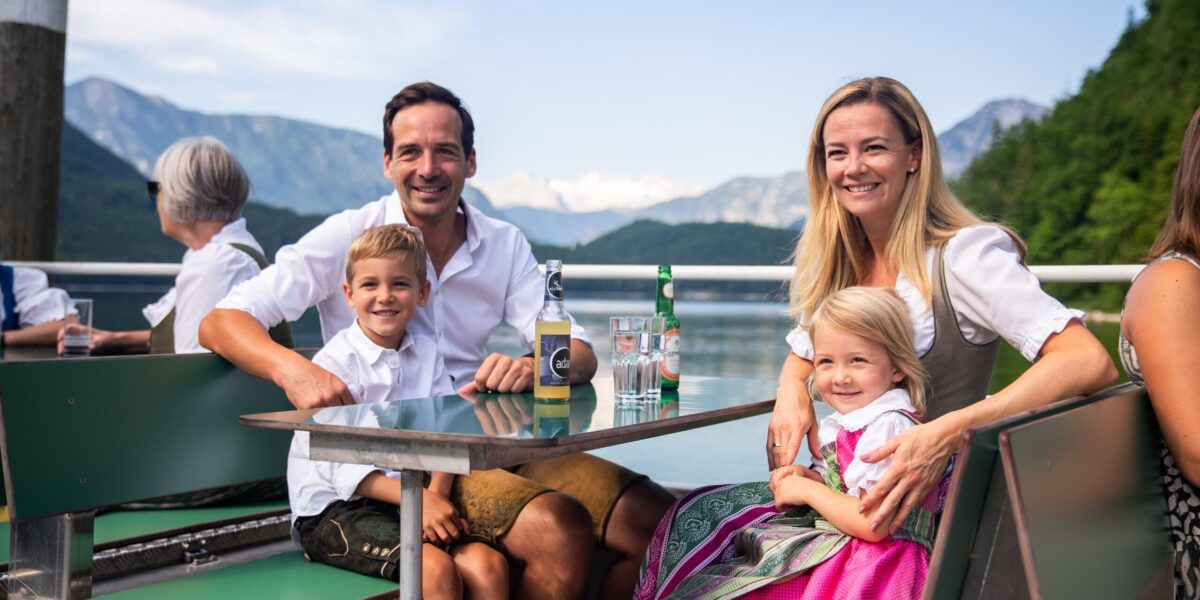Eine lächelnde Familie in traditioneller bayerischer Kleidung sitzt an einem Tisch im Freien an einem See mit Bergen im Hintergrund.