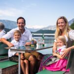 Eine lächelnde Familie in traditioneller bayerischer Kleidung sitzt an einem Tisch im Freien an einem See mit Bergen im Hintergrund.