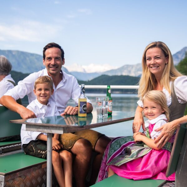 Eine lächelnde Familie in traditioneller bayerischer Kleidung sitzt an einem Tisch im Freien an einem See mit Bergen im Hintergrund.