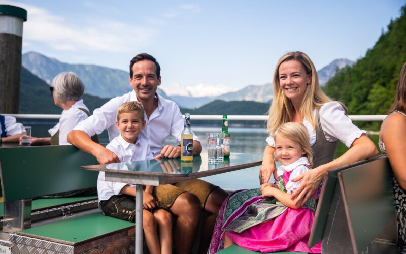 Eine lächelnde Familie in traditioneller bayerischer Kleidung sitzt an einem Tisch im Freien an einem See mit Bergen im Hintergrund.