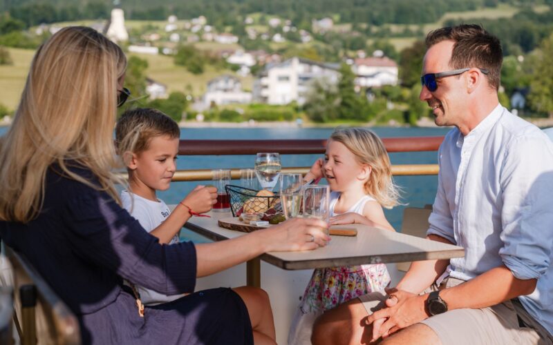 Eine vierköpfige Familie sitzt an einem sonnigen Tag an einem Tisch im Freien und genießt Getränke und Snacks vor einer malerischen Landschaft im Hintergrund.