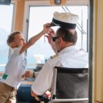 Ein Kind greift spielerisch nach einer Kapitänsmütze und setzt sie einem Erwachsenen auf, der am Steuer eines Bootes sitzt.