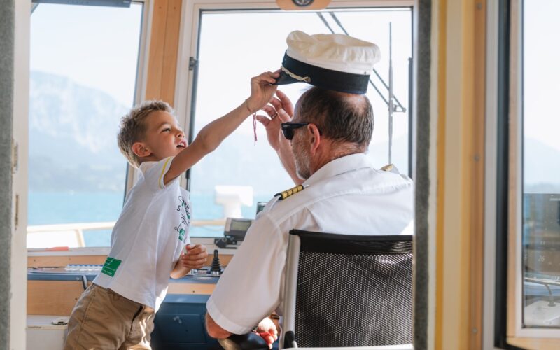 Ein Kind greift spielerisch nach einer Kapitänsmütze und setzt sie einem Erwachsenen auf, der am Steuer eines Bootes sitzt.