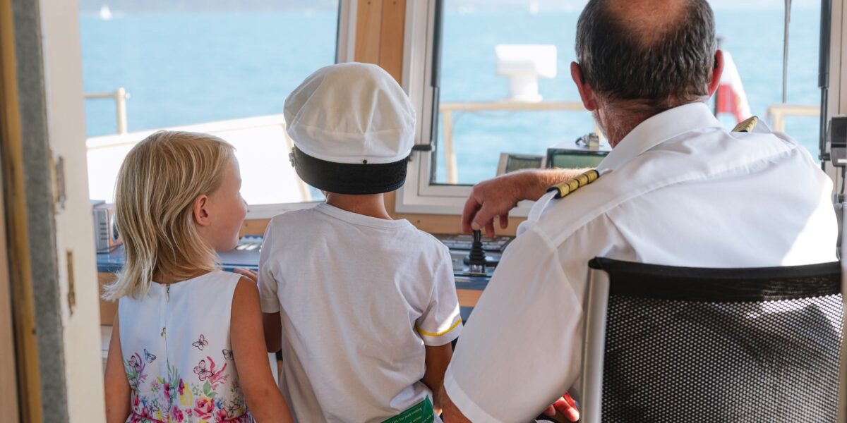 Ein Schiffskapitän sitzt mit zwei kleinen Kindern, von denen eines eine Kapitänsmütze trägt, am Steuerpult. Sie schauen durch das Kabinenfenster auf das Wasser hinaus.