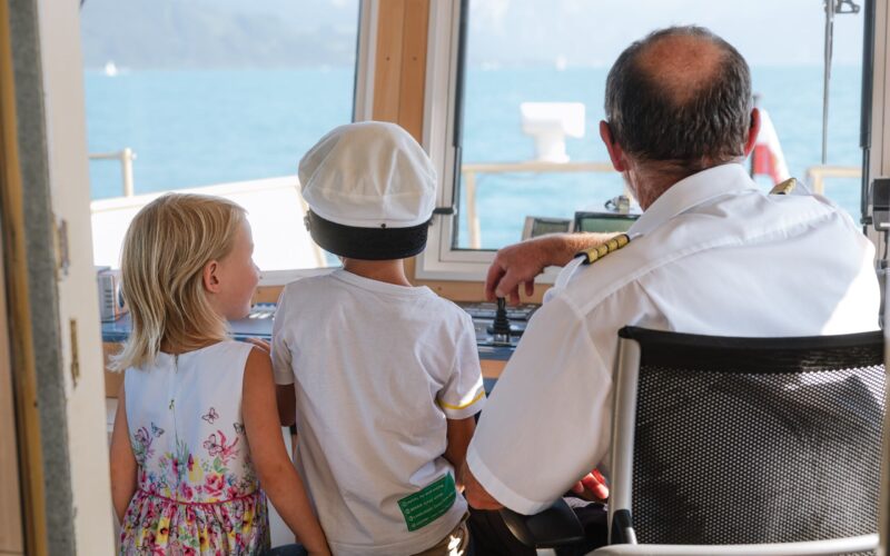 Ein Schiffskapitän sitzt mit zwei kleinen Kindern, von denen eines eine Kapitänsmütze trägt, am Steuerpult. Sie schauen durch das Kabinenfenster auf das Wasser hinaus.