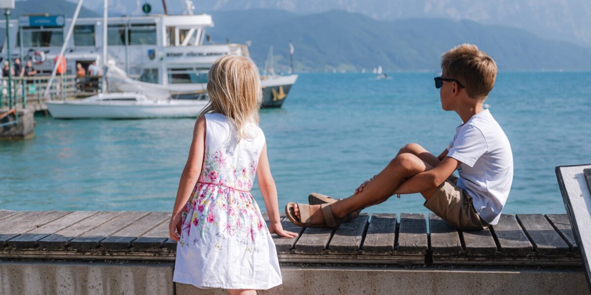 Ein Mädchen in einem weißen Kleid und ein Junge mit Sonnenbrille sitzen an einem hölzernen Steg mit Blick auf Boote auf einem ruhigen See mit Bergen im Hintergrund.