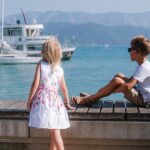 Ein Mädchen in einem weißen Kleid und ein Junge mit Sonnenbrille sitzen an einem hölzernen Steg mit Blick auf Boote auf einem ruhigen See mit Bergen im Hintergrund.