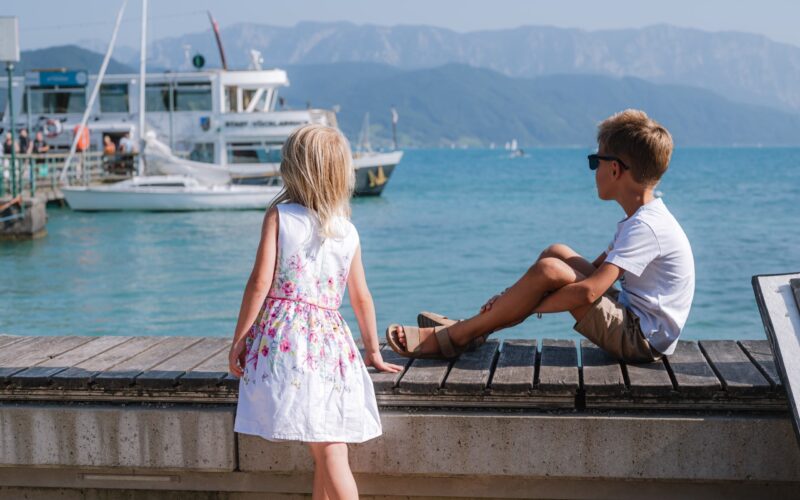 Ein Mädchen in einem weißen Kleid und ein Junge mit Sonnenbrille sitzen an einem hölzernen Steg mit Blick auf Boote auf einem ruhigen See mit Bergen im Hintergrund.