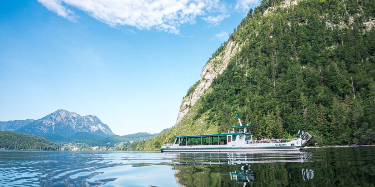 Ein weißes Ausflugsboot fährt über einen ruhigen See, der von bewaldeten Hügeln und Bergen umgeben ist, unter einem blauen Himmel mit vereinzelten Wolken.