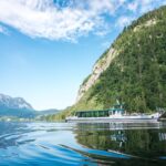 Ein weißes Ausflugsboot fährt über einen ruhigen See, der von bewaldeten Hügeln und Bergen umgeben ist, unter einem blauen Himmel mit vereinzelten Wolken.