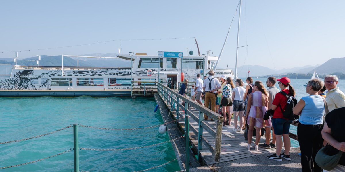 Eine Gruppe von Menschen wartet an einem klaren, sonnigen Tag am türkisblauen Wasser in einer Schlange an einem Dock, um an Bord eines weißen Passagierschiffs zu gehen.