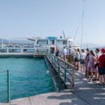 Eine Gruppe von Menschen wartet an einem klaren, sonnigen Tag am türkisblauen Wasser in einer Schlange an einem Dock, um an Bord eines weißen Passagierschiffs zu gehen.