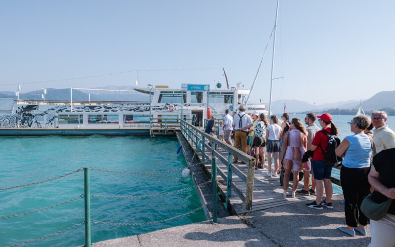 Eine Gruppe von Menschen wartet an einem klaren, sonnigen Tag am türkisblauen Wasser in einer Schlange an einem Dock, um an Bord eines weißen Passagierschiffs zu gehen.