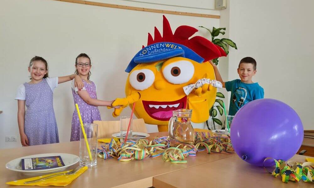 Drei Kinder lächeln und posieren mit einem großen gelben Maskottchen in einem Raum, der mit bunten Partyartikeln und einem lila Luftballon auf einem Tisch dekoriert ist.