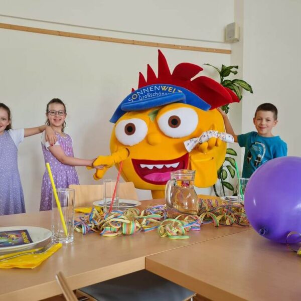 Drei Kinder lächeln und posieren mit einem großen gelben Maskottchen in einem Raum, der mit bunten Partyartikeln und einem lila Luftballon auf einem Tisch dekoriert ist.