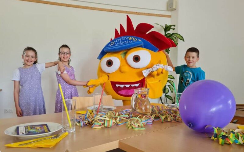 Drei Kinder lächeln und posieren mit einem großen gelben Maskottchen in einem Raum, der mit bunten Partyartikeln und einem lila Luftballon auf einem Tisch dekoriert ist.