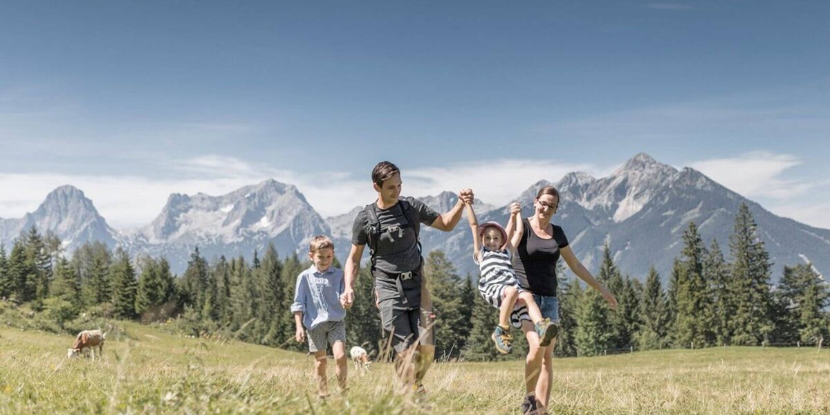 Eine vierköpfige Familie geht auf einer Wiese mit Bergen im Hintergrund spazieren; zwei Erwachsene halten sich an den Händen, ein Kind schaukelt zwischen ihnen, während ein anderes Kind neben ihnen läuft.