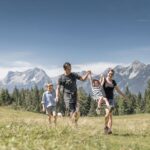 Eine vierköpfige Familie geht auf einer Wiese mit Bergen im Hintergrund spazieren; zwei Erwachsene halten sich an den Händen, ein Kind schaukelt zwischen ihnen, während ein anderes Kind neben ihnen läuft.