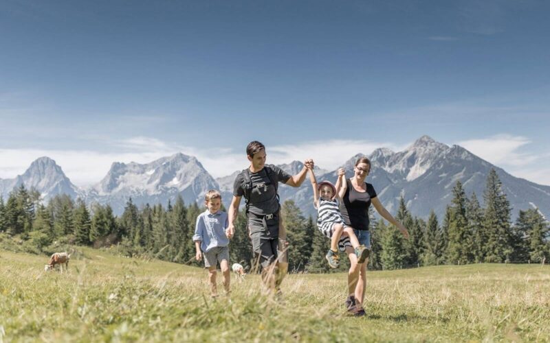 Eine vierköpfige Familie geht auf einer Wiese mit Bergen im Hintergrund spazieren; zwei Erwachsene halten sich an den Händen, ein Kind schaukelt zwischen ihnen, während ein anderes Kind neben ihnen läuft.