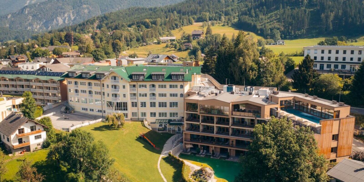 Luftaufnahme eines modernen Hotelkomplexes mit mehreren Gebäuden, umgeben von grünen Rasenflächen, Bäumen und Bergen im Hintergrund.