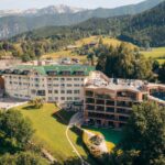 Luftaufnahme eines modernen Hotelkomplexes mit mehreren Gebäuden, umgeben von grünen Rasenflächen, Bäumen und Bergen im Hintergrund.