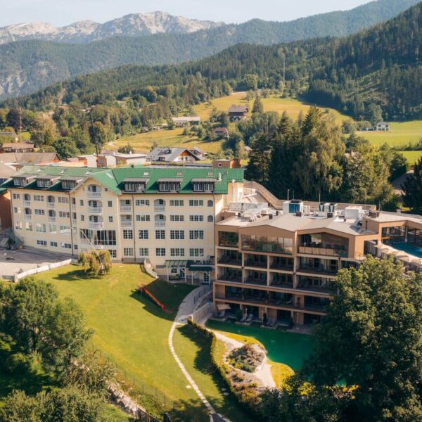 Luftaufnahme eines modernen Hotelkomplexes mit mehreren Gebäuden, umgeben von grünen Rasenflächen, Bäumen und Bergen im Hintergrund.