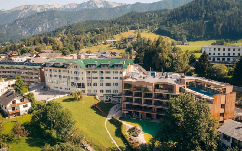 Luftaufnahme eines modernen Hotelkomplexes mit mehreren Gebäuden, umgeben von grünen Rasenflächen, Bäumen und Bergen im Hintergrund.