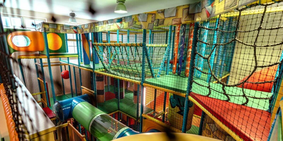 Indoor-Spielplatz mit Kletternetzen, Rutschen, bunten Tunneln und gepolsterten Spielstrukturen unter Leuchtstoffröhren.