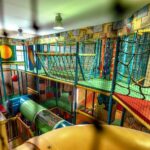 Indoor-Spielplatz mit Kletternetzen, Rutschen, bunten Tunneln und gepolsterten Spielstrukturen unter Leuchtstoffröhren.