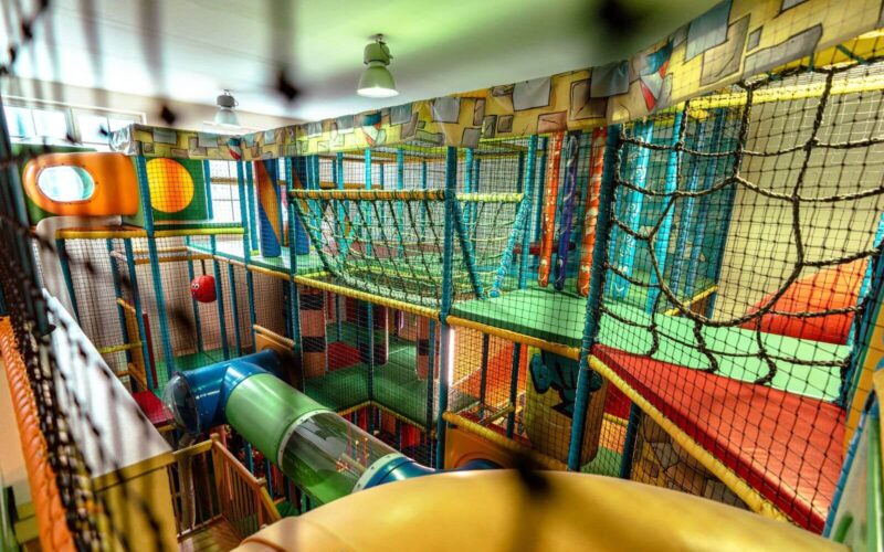 Indoor-Spielplatz mit Kletternetzen, Rutschen, bunten Tunneln und gepolsterten Spielstrukturen unter Leuchtstoffröhren.