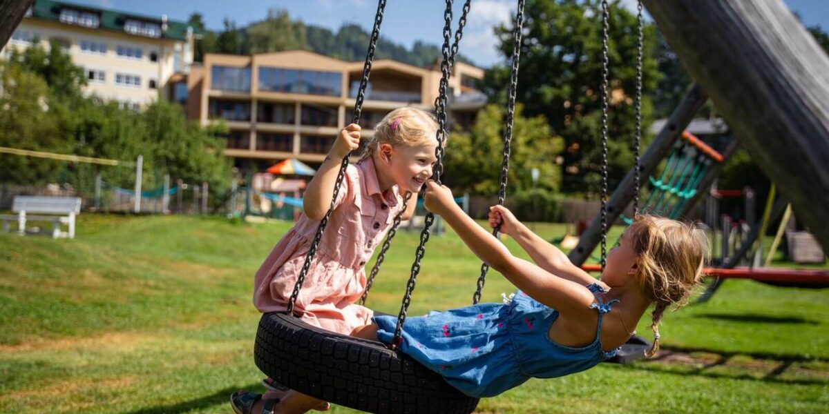 Zwei junge Mädchen in Kleidern spielen und halten sich an den Händen, während sie an einem sonnigen Tag auf einer Reifenschaukel auf einem grasbewachsenen Spielplatz sitzen.