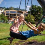 Zwei junge Mädchen in Kleidern spielen und halten sich an den Händen, während sie an einem sonnigen Tag auf einer Reifenschaukel auf einem grasbewachsenen Spielplatz sitzen.
