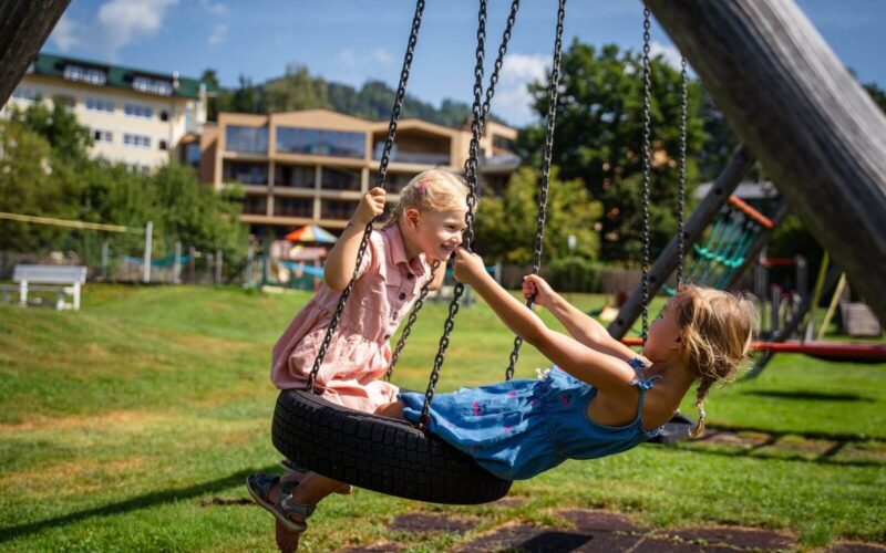 Zwei junge Mädchen in Kleidern spielen und halten sich an den Händen, während sie an einem sonnigen Tag auf einer Reifenschaukel auf einem grasbewachsenen Spielplatz sitzen.