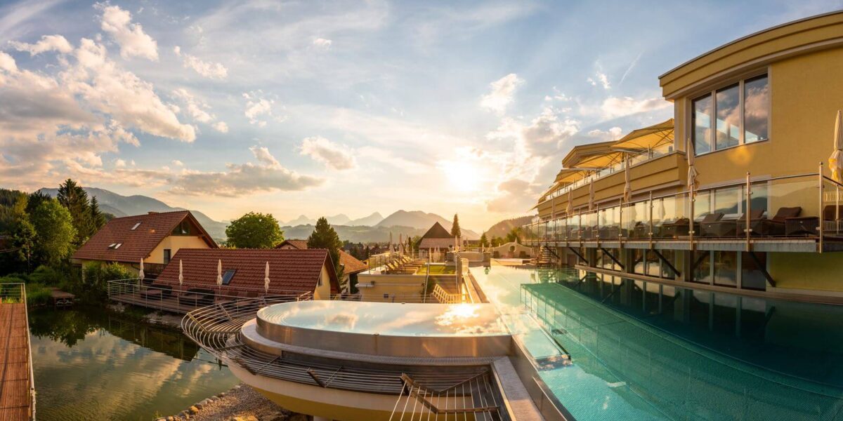 Modernes Gebäude mit einem Infinity-Pool mit Blick auf einen Teich, die Dächer und die fernen Berge bei Sonnenuntergang unter einem teilweise bewölkten Himmel.
