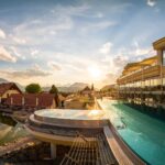 Modernes Gebäude mit einem Infinity-Pool mit Blick auf einen Teich, die Dächer und die fernen Berge bei Sonnenuntergang unter einem teilweise bewölkten Himmel.